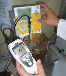 Ein elektrisches Thermometer mit Messfühler in einem Temperaturbad sowie einer Hand im Hintergrund an gelb-weißer Steuereinheit des Temperaturbades