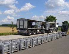 Ein Eichfahrzeug mit Anhänger steht auf einer Fahrzeugwaage, vor der Waage stehen neun Gewichtskäfige mit je zwei 500 Kilogramm-Rollengewichten aus Stahl, ein Gewichtskäfig wird gerade von einem Gabelstapler angehoben