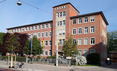 rotes Backsteingebäude vor blauem Himmel, mit Bäumen und Fahnen davor
