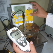 Ein elektrisches Thermometer mit Messfühler in einem Temperaturbad sowie einer Hand im Hintergrund an gelb-weißer Steuereinheit des Temperaturbades
