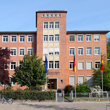 Ein rotes fünfstöckiges Backsteingebäude vor blauem Himmel mit wehenden Fahnen im Vordergrund