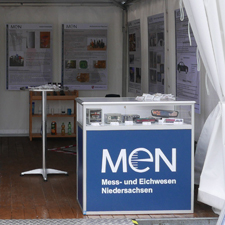 Vitrine mit blauem Hintergrund darauf MEN, daneben ein Stehtisch und aufgehängte Bilder an Zeltwand