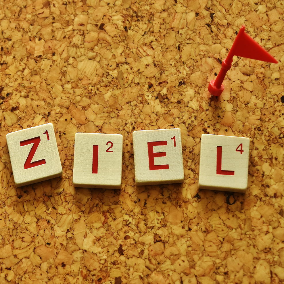 4 weiße Würfel mit Text ZIEL auf einer Korkpinnwand mit einem roten Fähnchen-Stecknadel