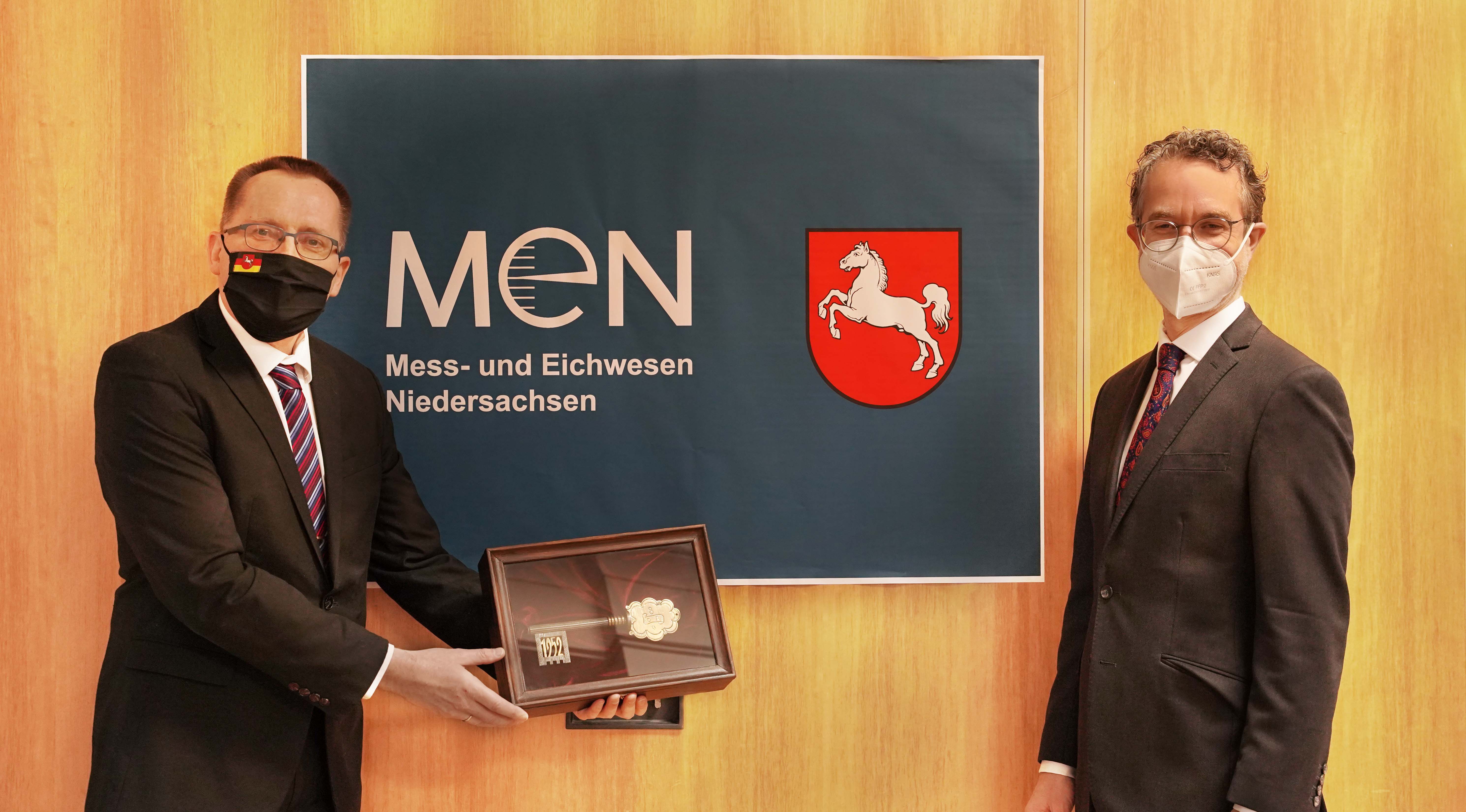 2 Männer mit Masken stehen vor dem Logo des Mess -und Eichwesen auf dem das Niedersachsenpferd zu sehen ist. Der linke Mann hält eine Kiste in dem ein goldener Schlüssel liegt hoch.