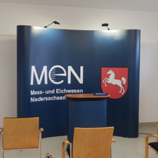 Eine blaue Messewand mit dem Niedersachsen-Logo und dem Logo des Mess-und Eichwesen Niedersachsen steht hinter einem blauen Rednerpult, davor stehen zwei Stühle